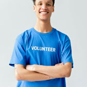 Young man in blue volunteer shirt smiling with arms crossed in a studio setting.