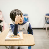 A cheerful child sitting at a desk in a classroom, holding a textbook.